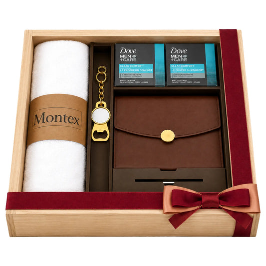 A men's grooming gift set in a light wood box with a dark red ribbon and bow. The set includes a white rolled towel with a brown paper band reading 'Montex', two black and blue boxes of Dove Men+Care Clean Comfort hand sanitizer, a brown leather wallet or card holder with a gold snap button, and a gold keychain bottle opener.