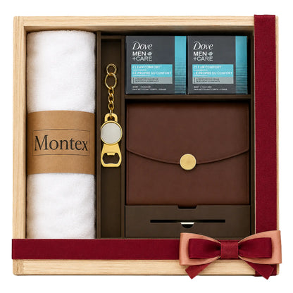 A Montex and Dove Men + Care gift set in a light wood box with a dark red ribbon and bow. The set includes a white rolled towel with a brown paper band reading 'Montex', two black and blue boxes of Dove Men + Care, a brown leather pouch with a gold snap, a gold keychain, and a bottle opener.