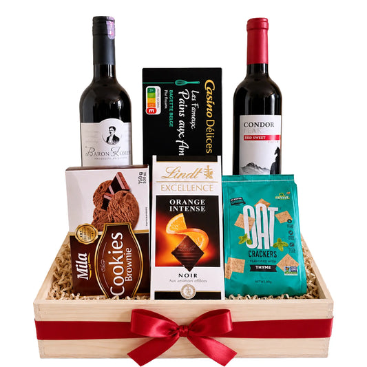 A wooden gift basket filled with assorted gourmet treats and beverages, tied with a red ribbon. The basket contains a bottle of Baron Louis red wine, a bottle of Condor red sweet wine, a box of Lindt Excellence orange intense chocolate, Mila Cookies Brownie, and a bag of Cat Bites Toffee Puffs, all arranged on shredded paper.