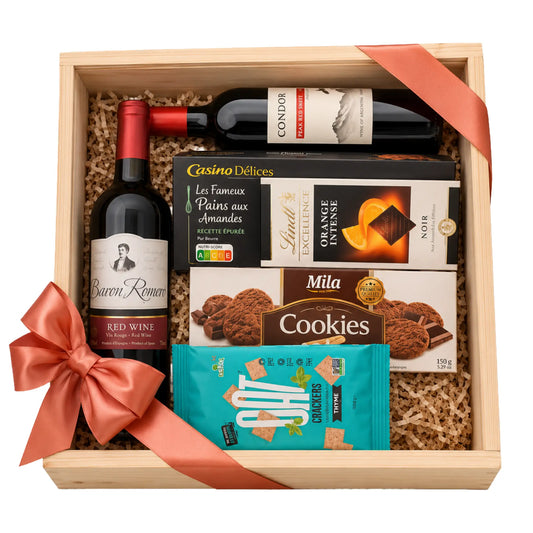 A wooden gift box filled with assorted gourmet treats and beverages, tied with a peach satin ribbon. The box contains a bottle of Baren Raymond red wine, a bottle of Condor cream wine, Casino Délices almond paste, Mila chocolate orange intense, and Choix Chocochip cookies.