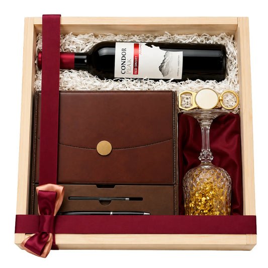 A wooden gift box filled with white crinkle paper and red satin ribbon, containing a bottle of Gondor Red Wine, a brown leather-bound journal with a gold snap button, a pen in a slot, and a clear textured wine glass with gold glitter inside red velvet.