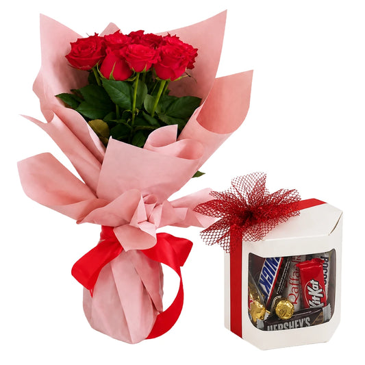 A gift set featuring a bouquet of six red roses wrapped in pink paper with a red ribbon, alongside a white gift box filled with assorted chocolates and wrapped candies, tied with a red mesh bow.