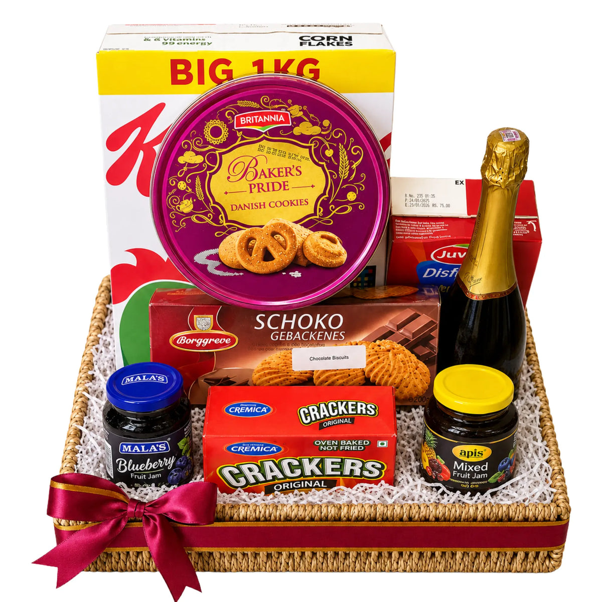 A curated gift basket arrangement in a woven tray with a red ribbon, featuring a large box of Kirana sweets, a round tin of Bikaneri Pride cookies, Schoko Gebakenes, blueberry and mixed fruit chutney jars, a box of Crackers, and a bottle of champagne on a white background.