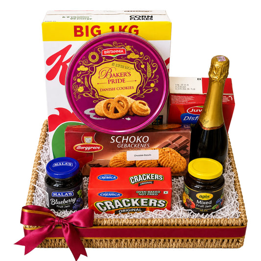 A curated gift basket arrangement in a woven tray with a red ribbon, featuring a large box of Kirana sweets, a round tin of Bikaneri Pride cookies, Schoko Gebakenes, blueberry and mixed fruit chutney jars, a box of Crackers, and a bottle of champagne on a white background.